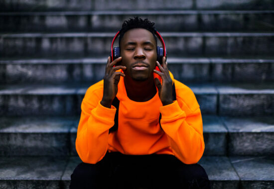 african-american man in stylish orange hoodie sweatshirt in wireless headphones listening music and enjoying music on background of stairs